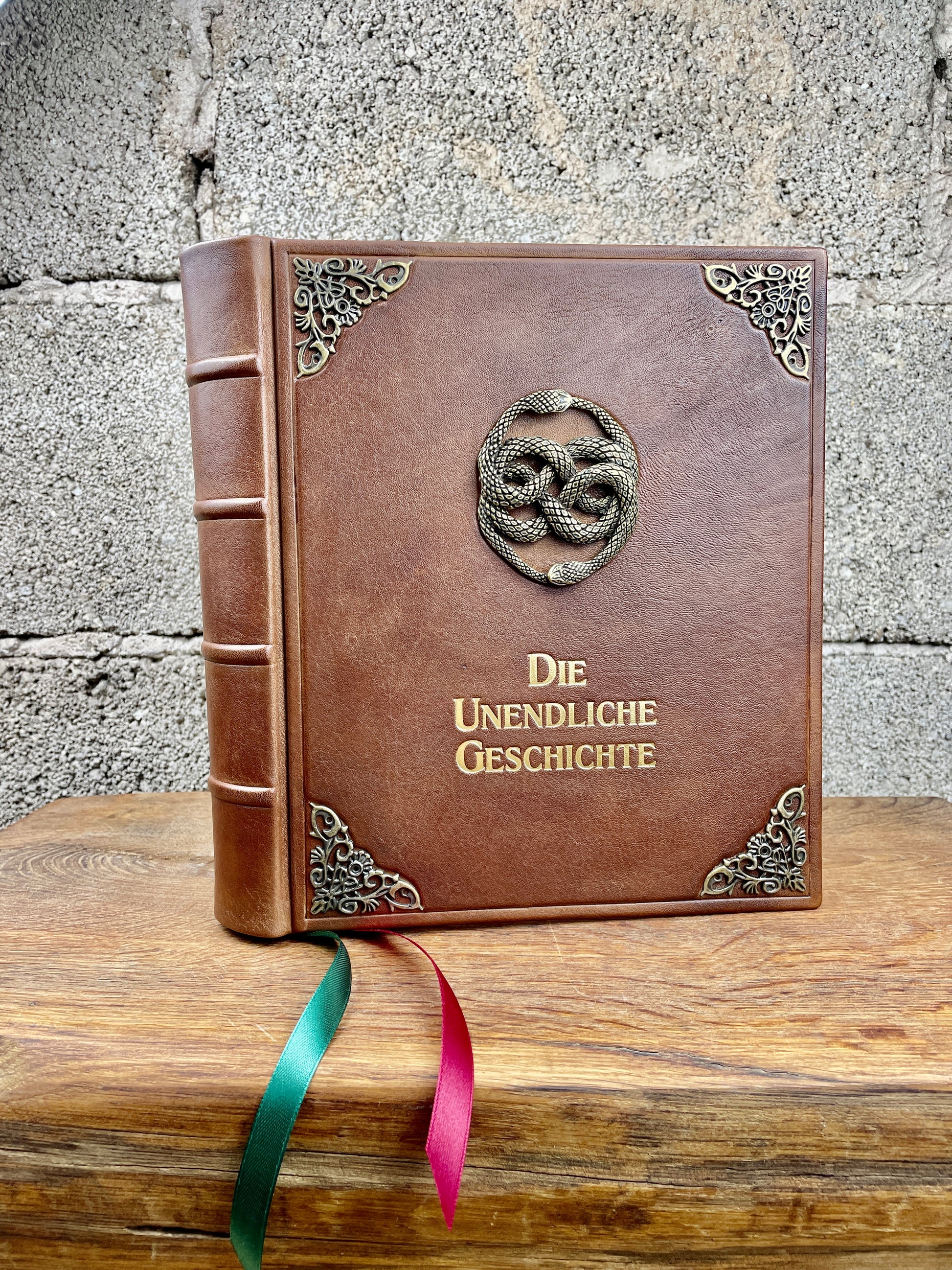 Lederausgabe der Unendlichen Geschichte mit großem AURYN-Emblem auf der Vorderseite, goldene Titelprägung, zwei breite Satin-Lesebänder in Rot und Grün, die luxuriös am Buch herabhängen.